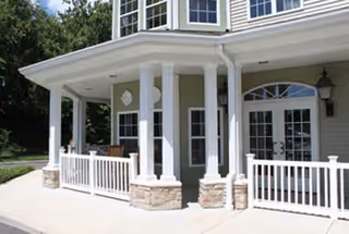Front porch entrance of a light-green building with white columns, railings, and stone accents.