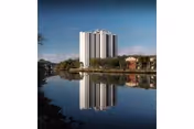 Tall apartment building reflected in a calm body of water with surrounding trees.