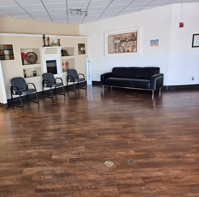 A spacious senior living lobby with four chairs along the wall, a black sofa, a fireplace display, and a Welcome sign.