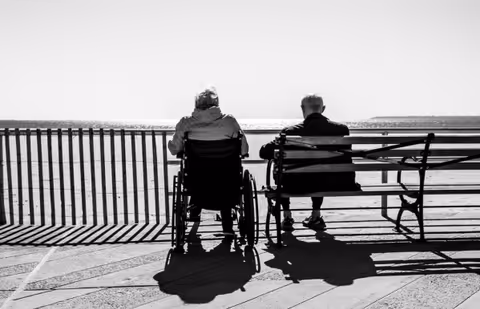 Two elderly people in wheelchairs sit on a bench beside a railing overlooking the water.