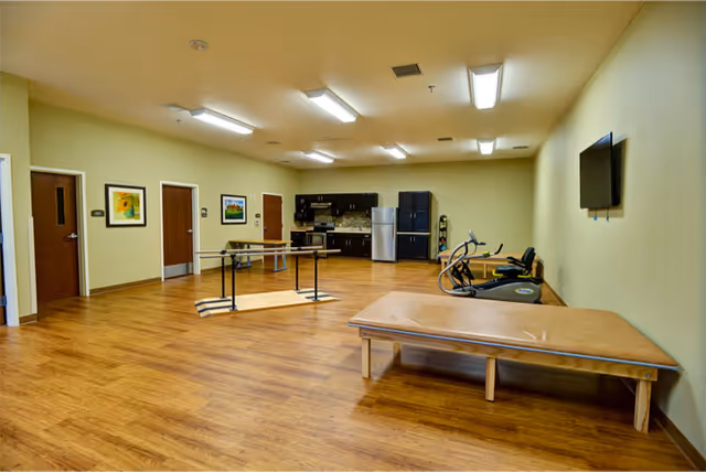 A spacious room with wooden flooring featuring exercise equipment including a stationary bike and parallel bars, a large padded table, a wall-mounted TV, and a kitchenette area with cabinets, a microwave, and a refrigerator. The room has several doors and framed artwork on the walls.