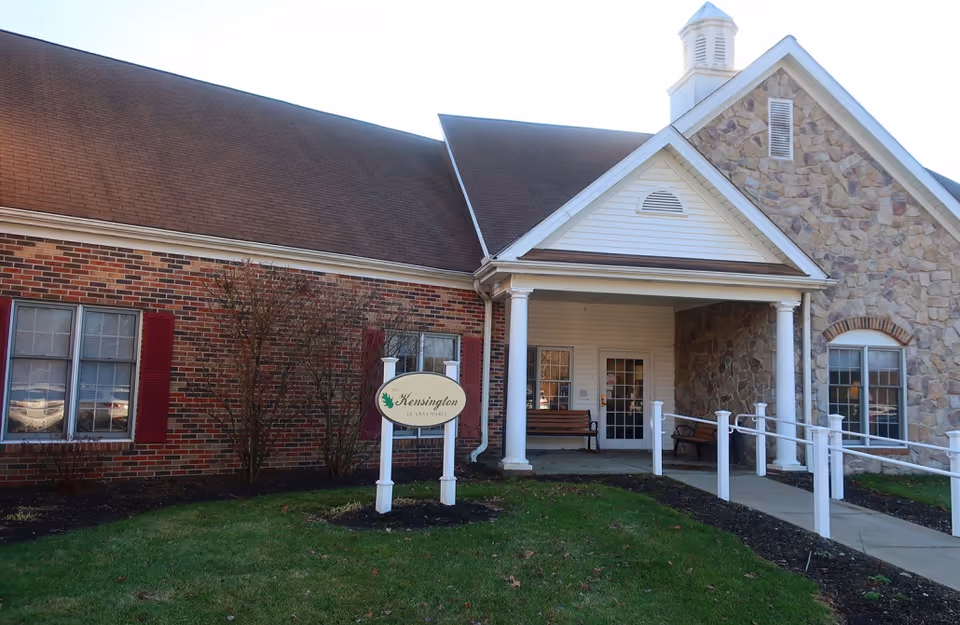 Front entrance of a brick-and-stone senior living facility with a covered porch, accessibility ramp, benches, and a 'Kensington' sign on the lawn.