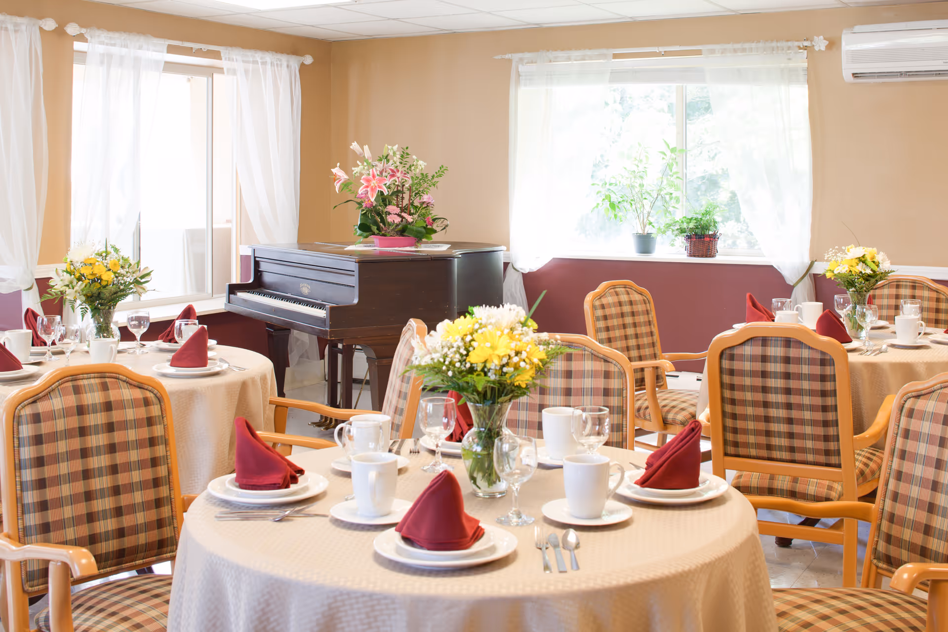A bright dining room with round tables covered in beige tablecloths, each set with white plates, cups, silverware, wine glasses, and red folded napkins. The chairs have a plaid pattern with wooden frames. There are flower arrangements on each table and a grand piano against the wall near two large windows with sheer white curtains. Potted plants are placed on the windowsills.