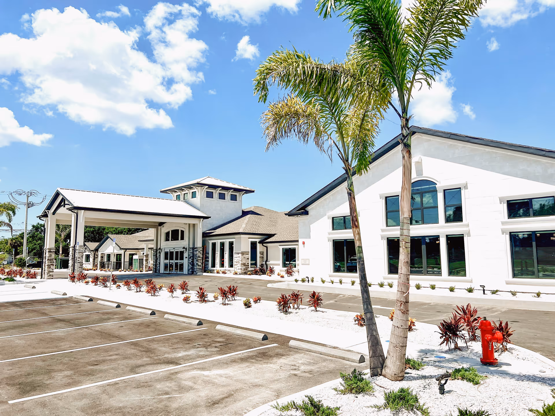 Exterior view of a modern senior living facility building with a covered entrance, palm trees, landscaped plants, and an empty parking lot under a partly cloudy blue sky.