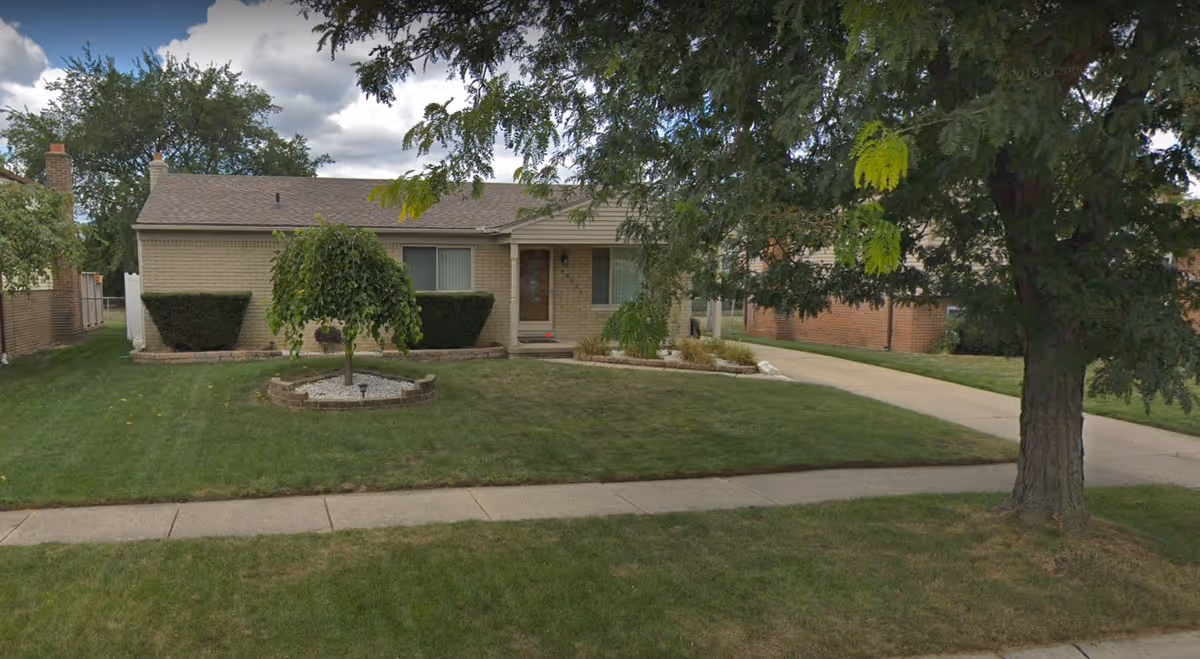 Single-story brick house with a small front porch, two windows, and a well-maintained lawn featuring a small tree surrounded by a circular stone border. A large tree is visible on the right side near the sidewalk and driveway.