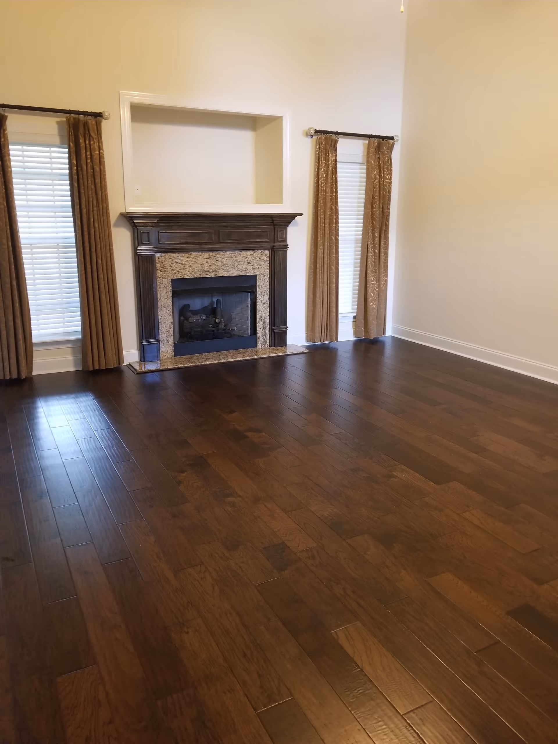 Bright living room featuring a stone fireplace, dark wood floor, and two brown curtains.