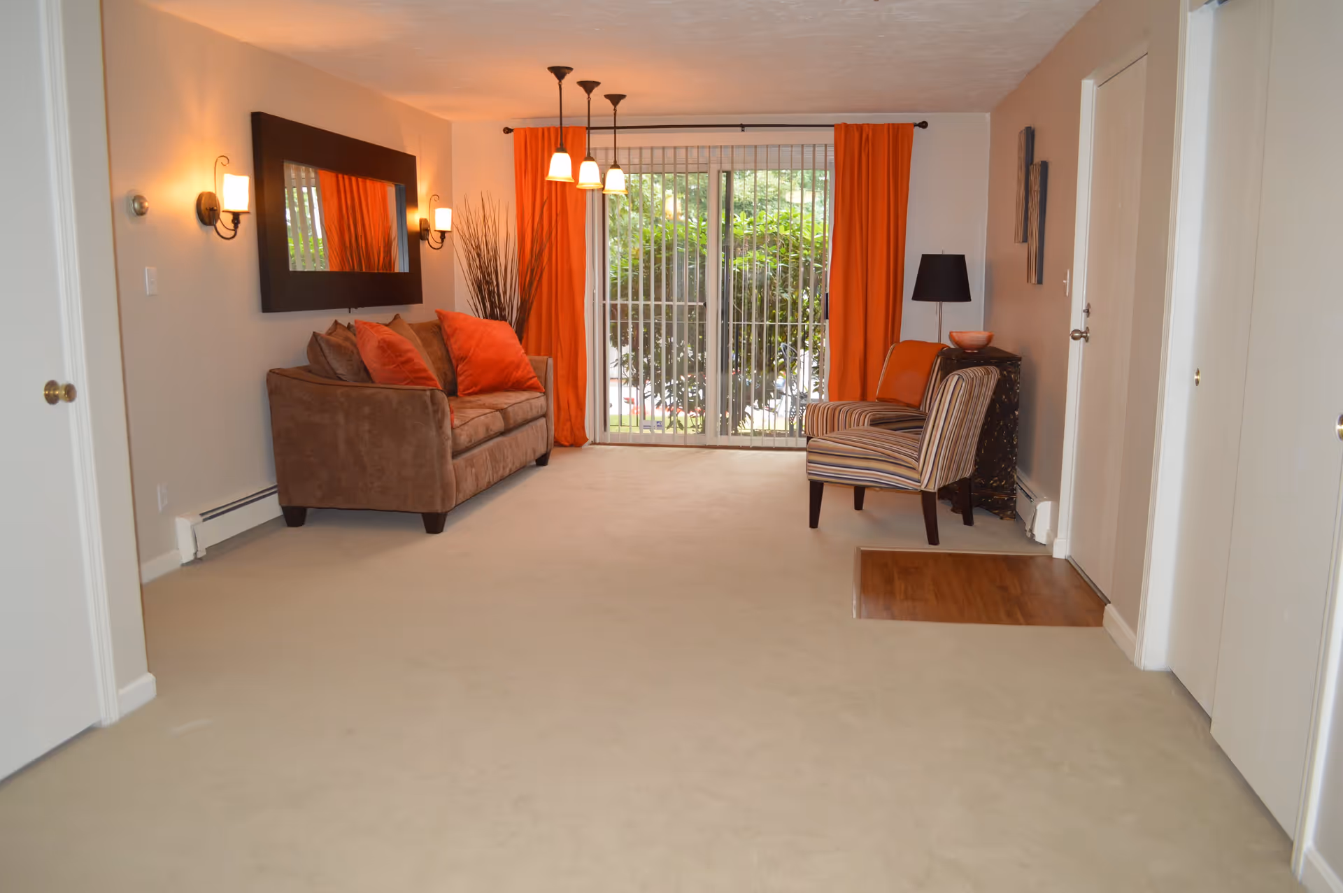 Spacious living room with a brown sofa and orange pillows, striped armchair, orange curtains, and a sliding glass door.