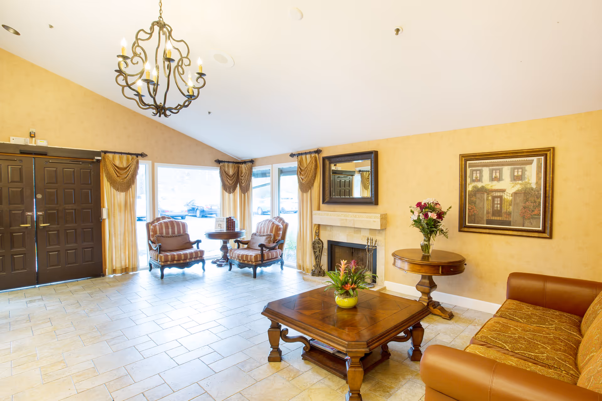A bright and spacious living room area with a high ceiling, featuring a chandelier, two striped armchairs with a small round table between them near large windows with curtains, a fireplace with a mirror above it, a wooden coffee table with a plant, a round side table with a vase of flowers, a framed painting on the wall, and a brown leather sofa.