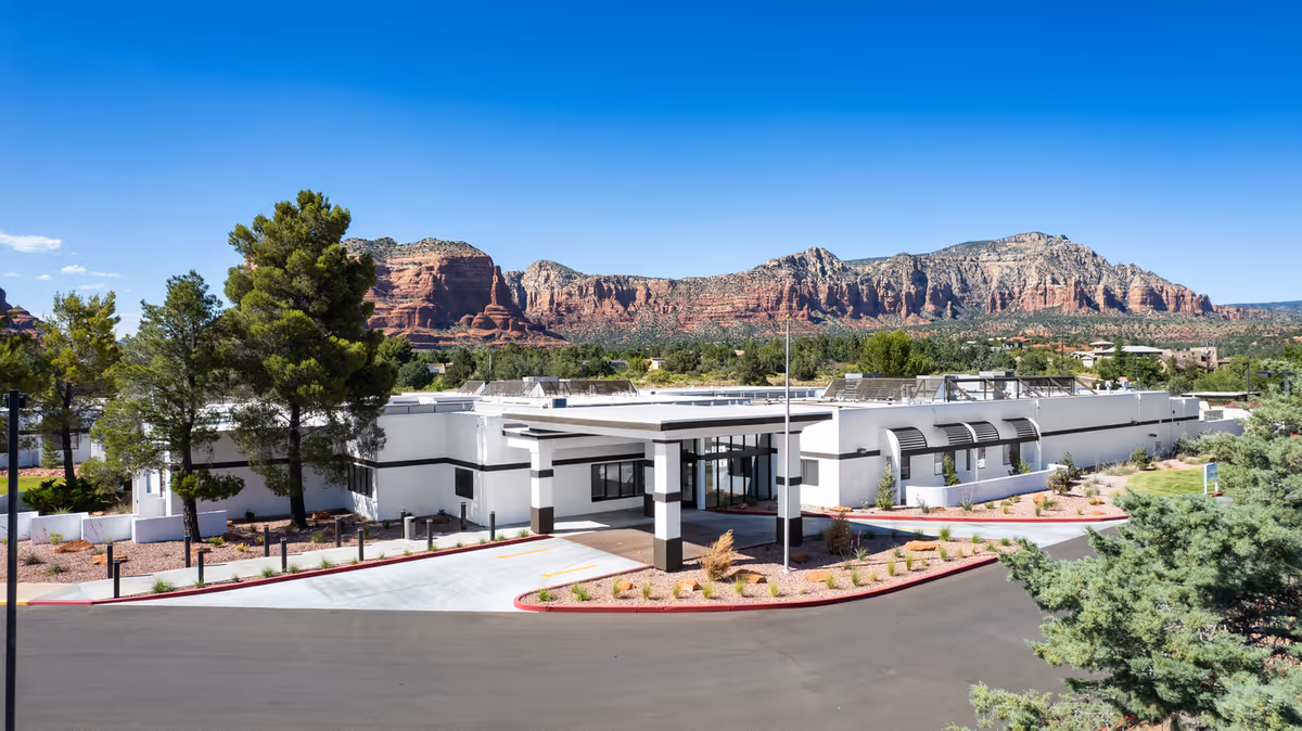 Exterior view of Haven Health Sedona facility with a modern white building surrounded by desert landscaping and trees, set against a backdrop of red rock mountains under a clear blue sky.