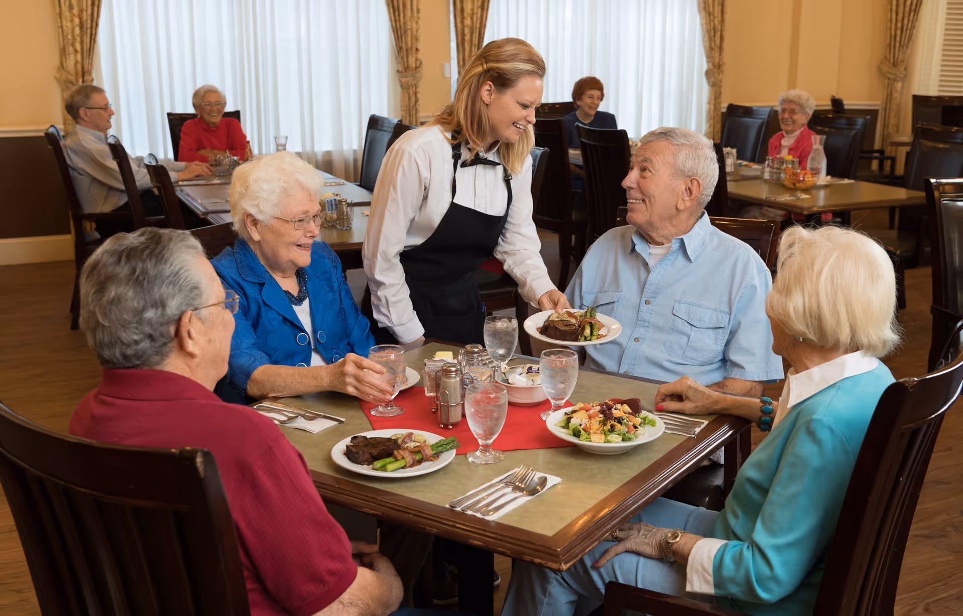 A group of elderly people sitting around a dining table in a retirement living facility, smiling and interacting with a waitress who is serving food. The table has plates with meals, glasses of water, and utensils. Other elderly people are seated at tables in the background.