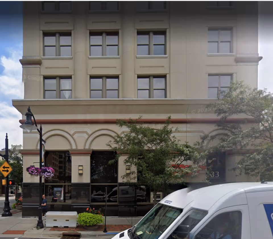 Beige multi-story building with arched storefronts on a city street, white van in foreground