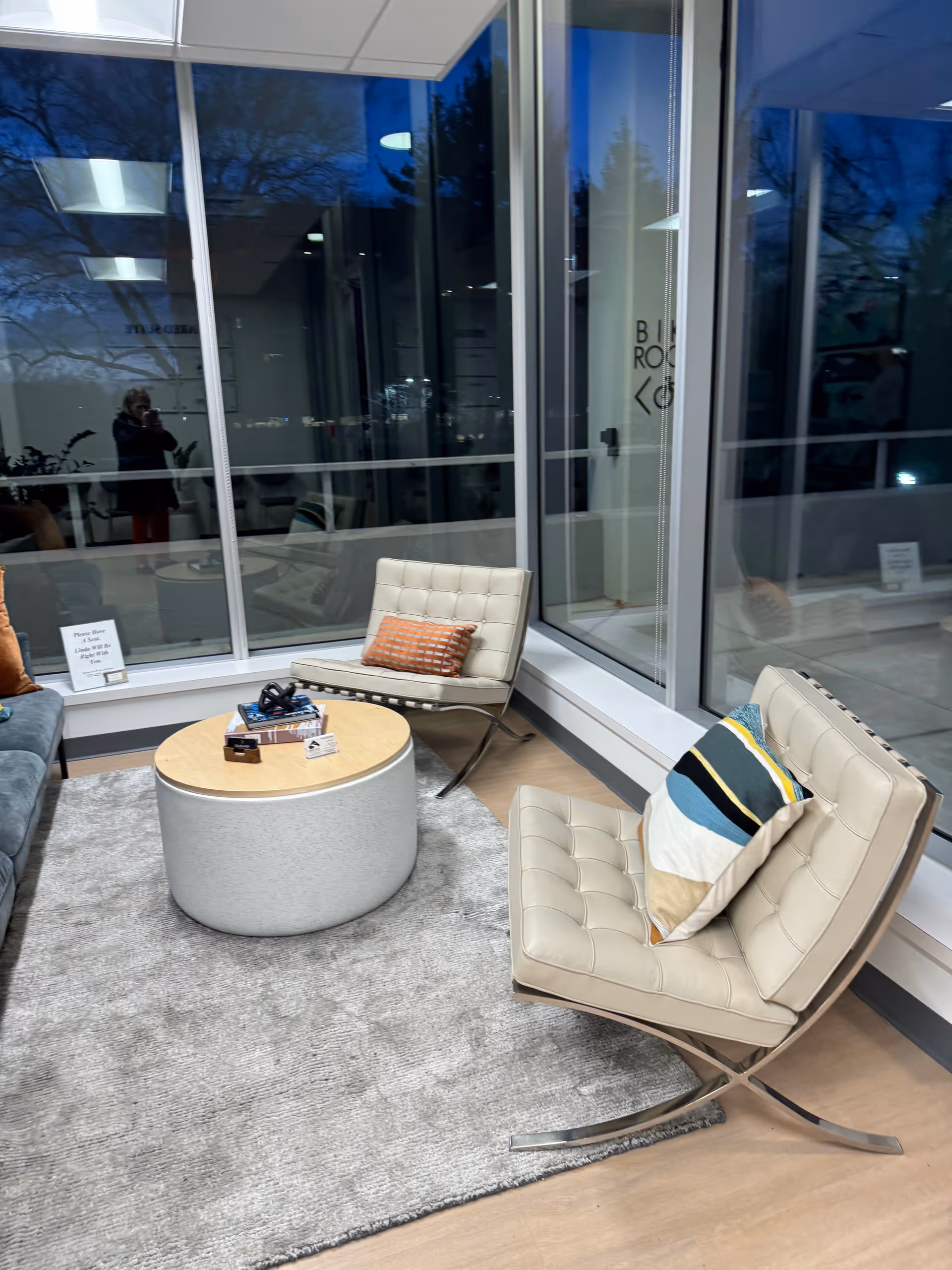 Modern indoor lounge with beige tufted chairs, a round coffee table, and glass windows.