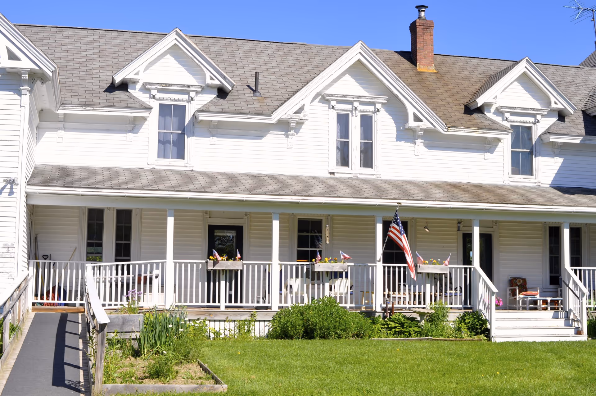 Front exterior view of a white two-story building with a porch, several windows, a chimney, an American flag, flower boxes on the porch railing, and a green lawn with garden beds in front.