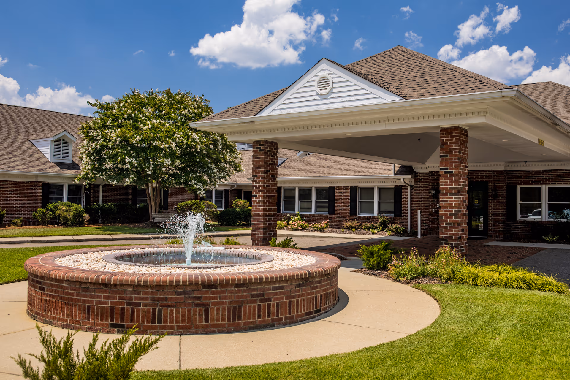Brick building with a circular fountain under a covered entrance and landscaped lawn.