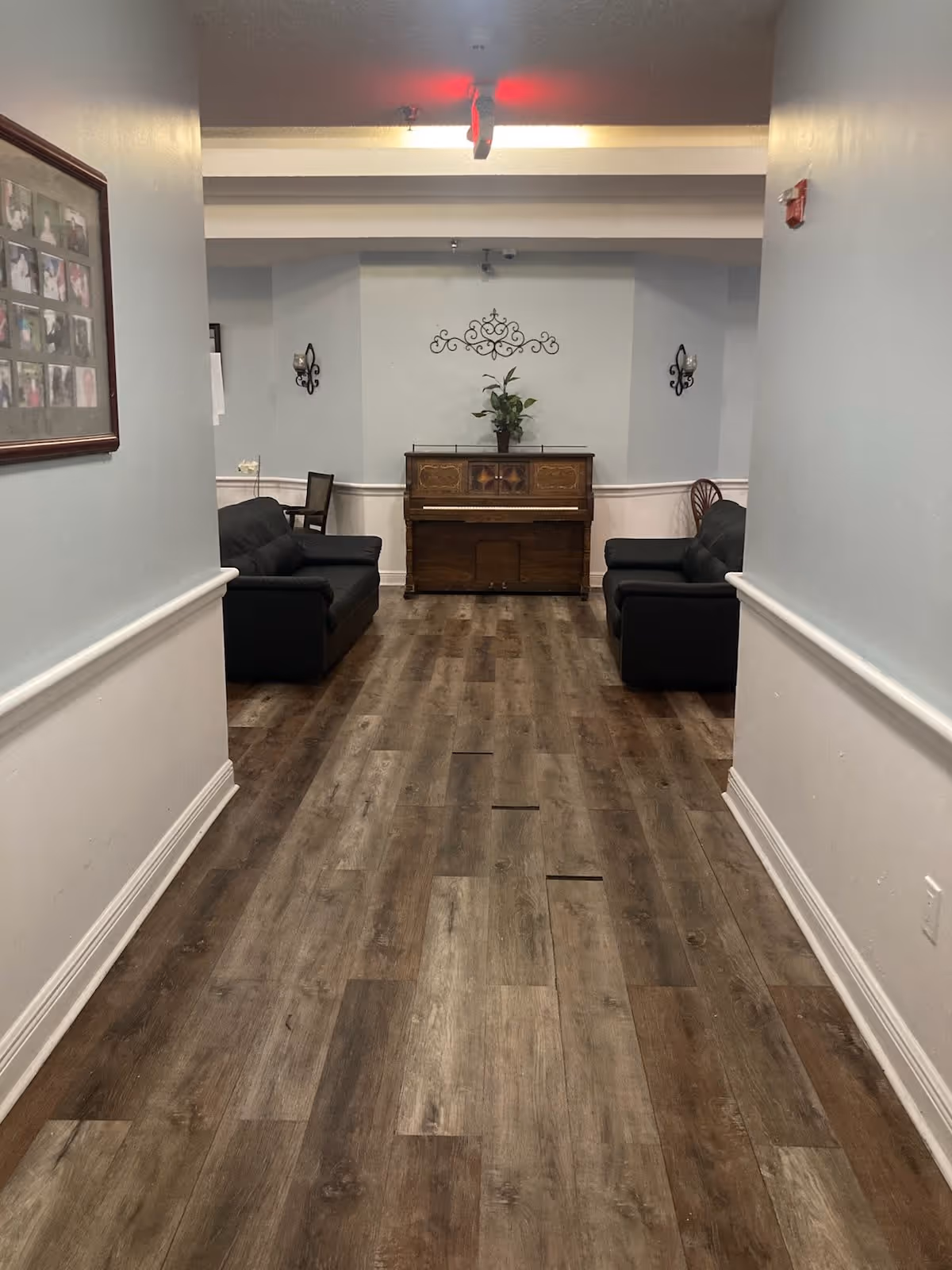 A hallway with wood flooring leading to a sitting area with two black sofas facing each other and a wooden piano against the far wall. There is a decorative metal wall piece above the piano and a potted plant on top of it. The walls are painted light blue with white wainscoting, and there is a framed photo collage on the left wall.