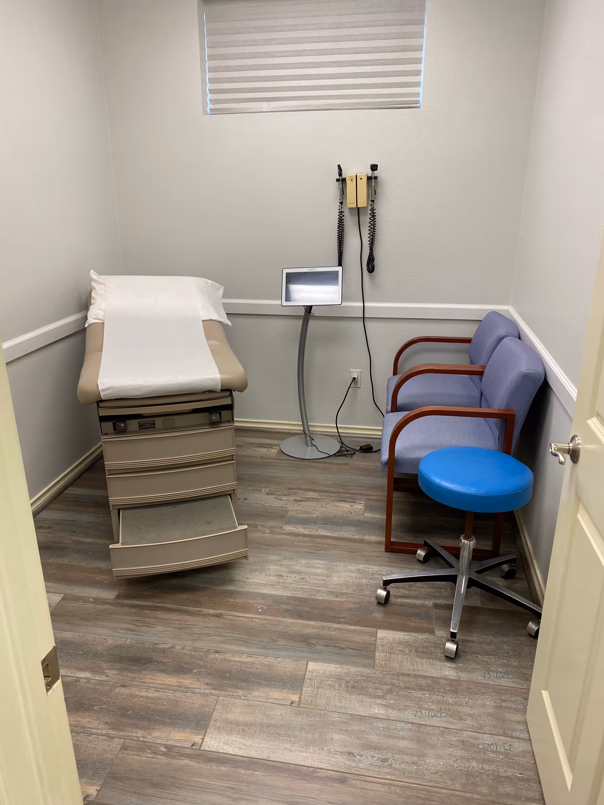 Small medical exam room with adjustable exam chair, wall monitor, blue stool, and blinds.