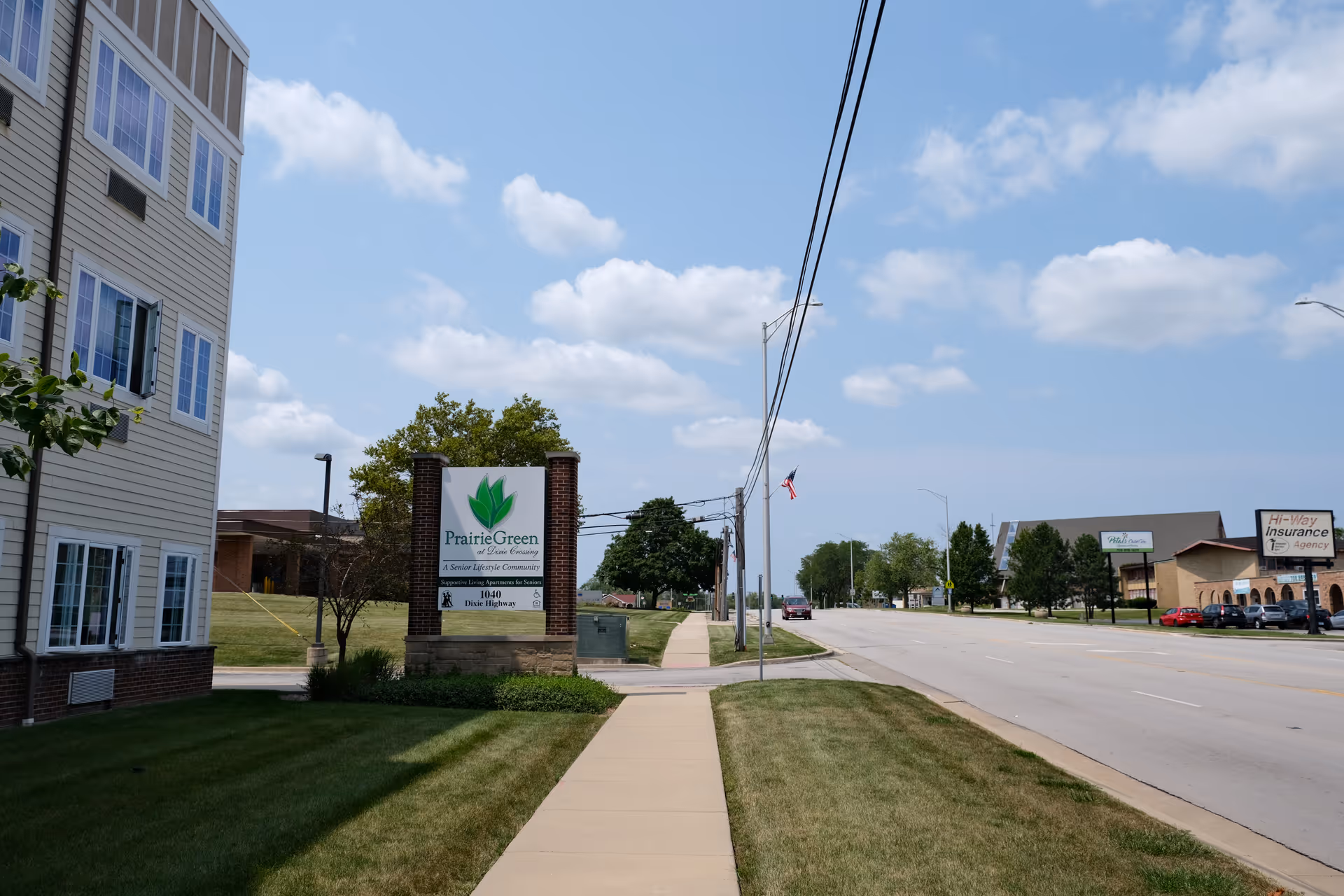 Sidewalk and lawn leading to a Prairie Green at Dixie Crossing sign beside a multi-story senior living building on a sunny street.