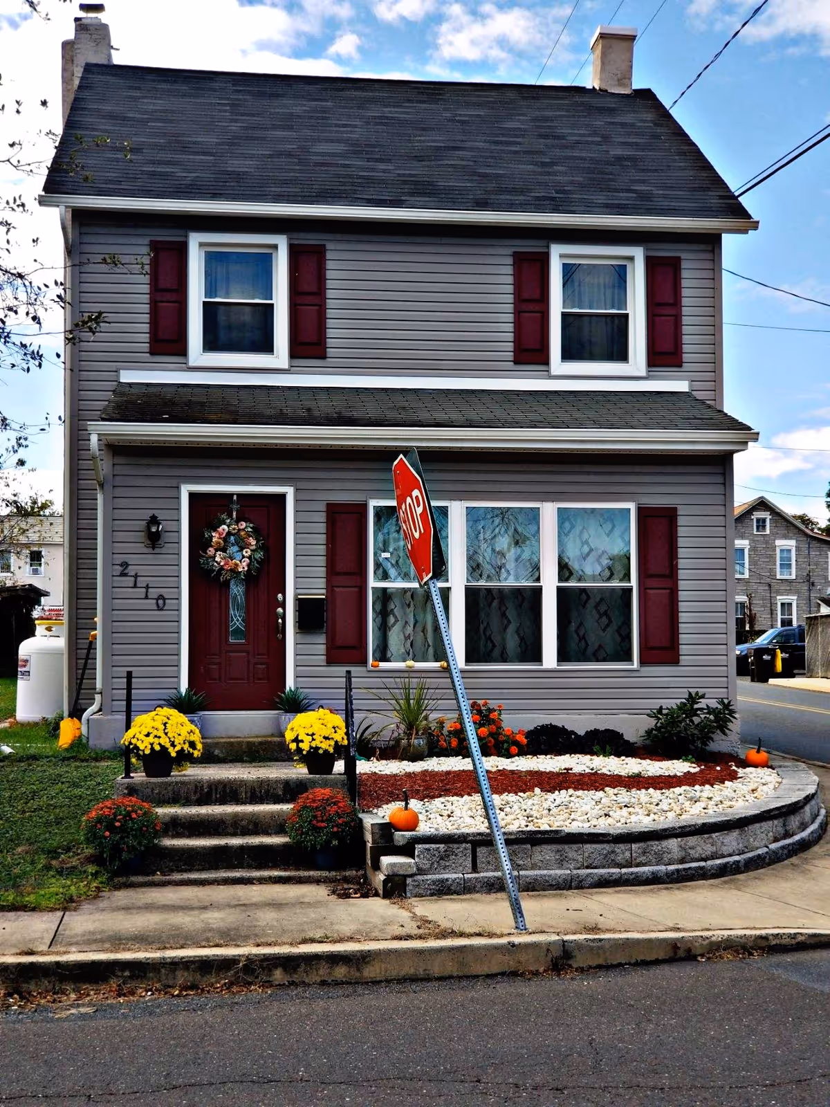 Two-story gray house with burgundy shutters and seasonal landscaping near a leaning stop sign.