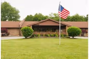 Single story brick senior living facility with flagpole, flag, and manicured lawn.