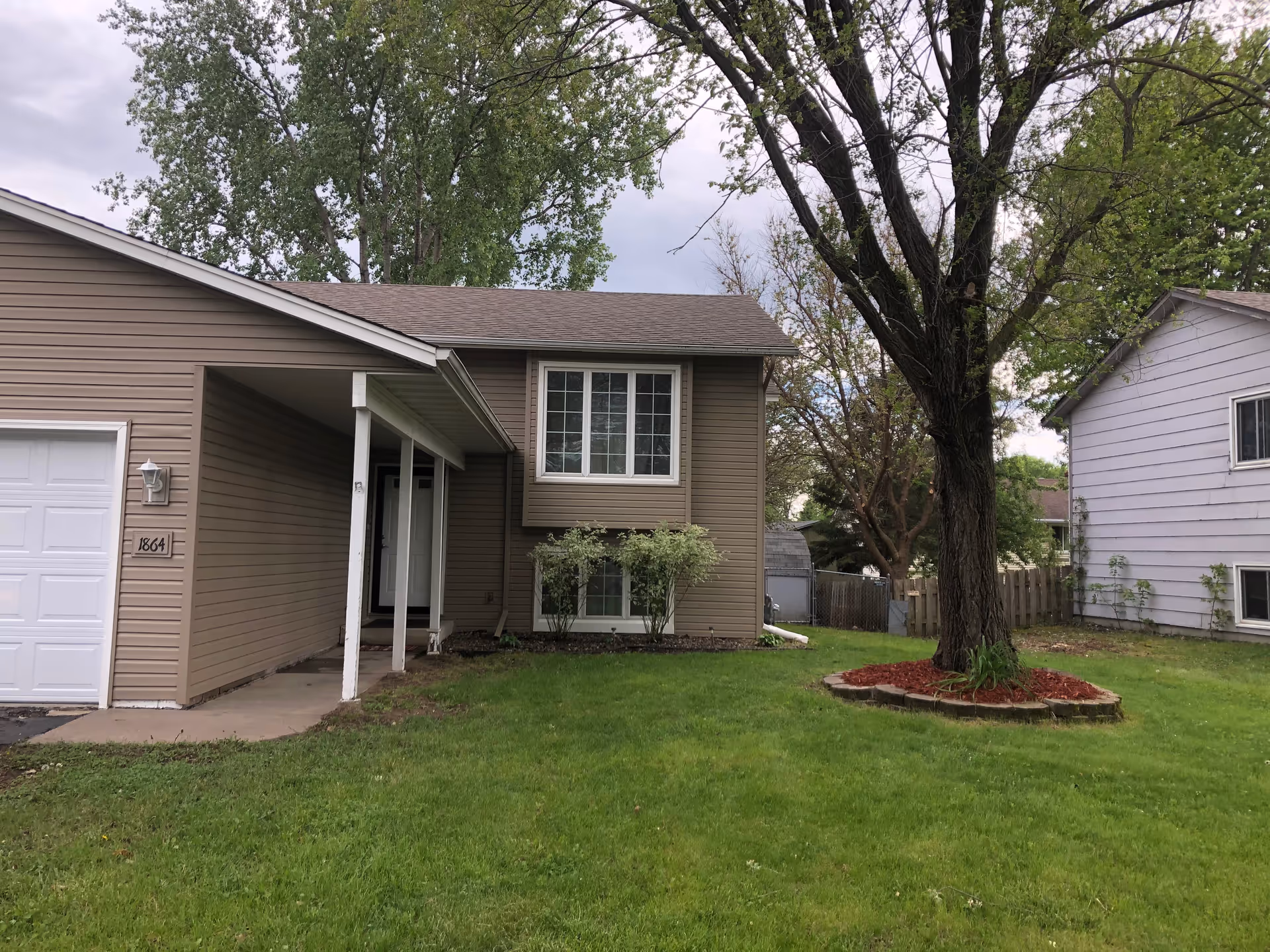 Two-story beige house with a front porch, attached garage, and a large tree in the yard.