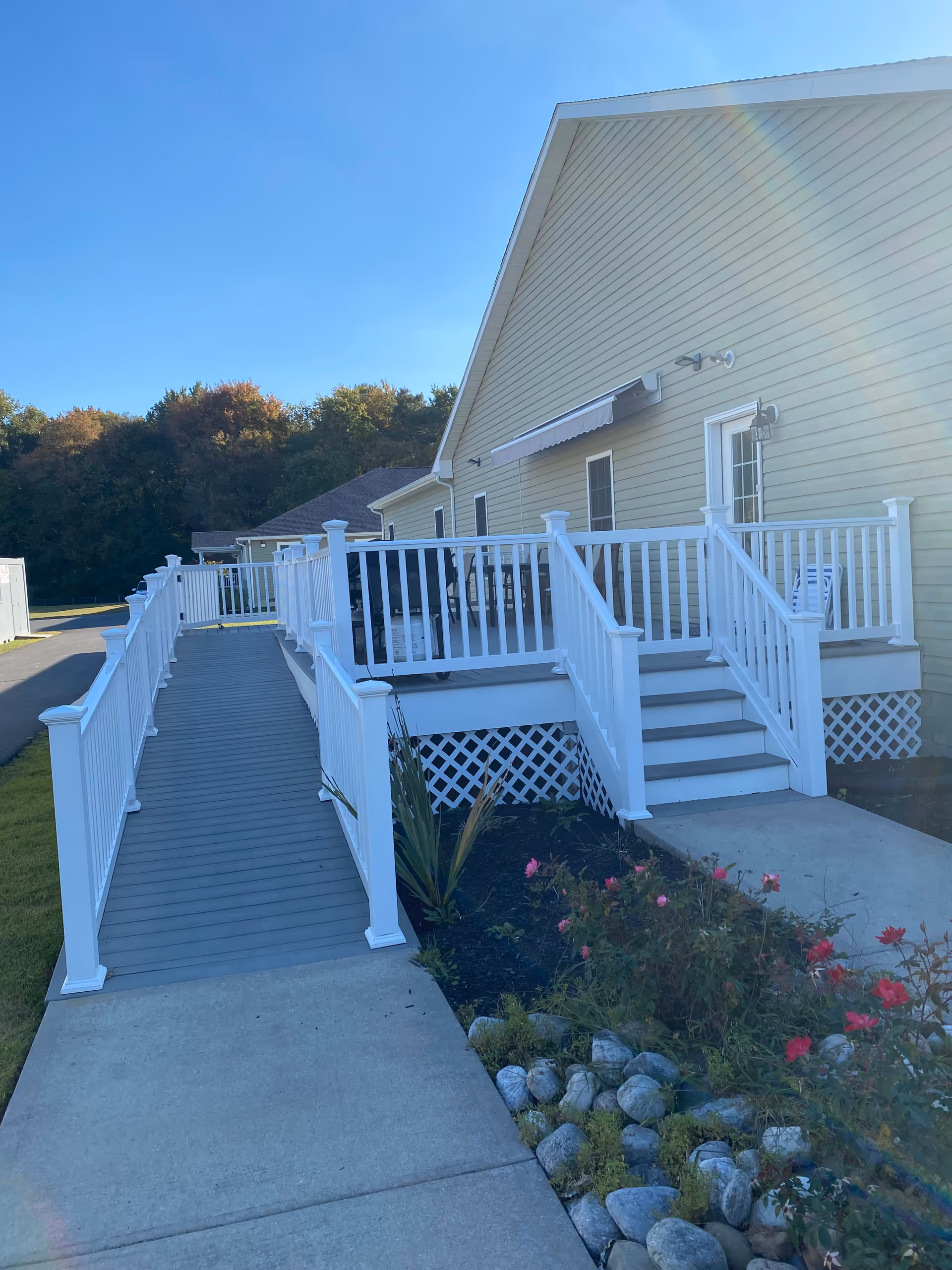 Exterior view of a light-yellow assisted living building with a white-railed accessibility ramp, deck, steps, and landscaped rocks and flowers.