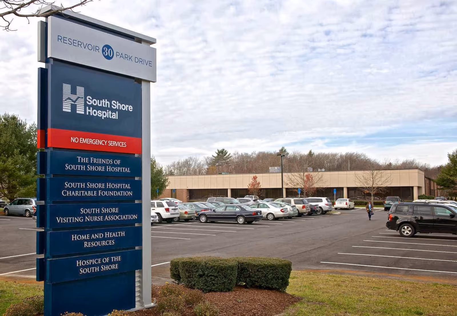 Tall sign for South Shore Hospital stands beside a parking lot and low building.