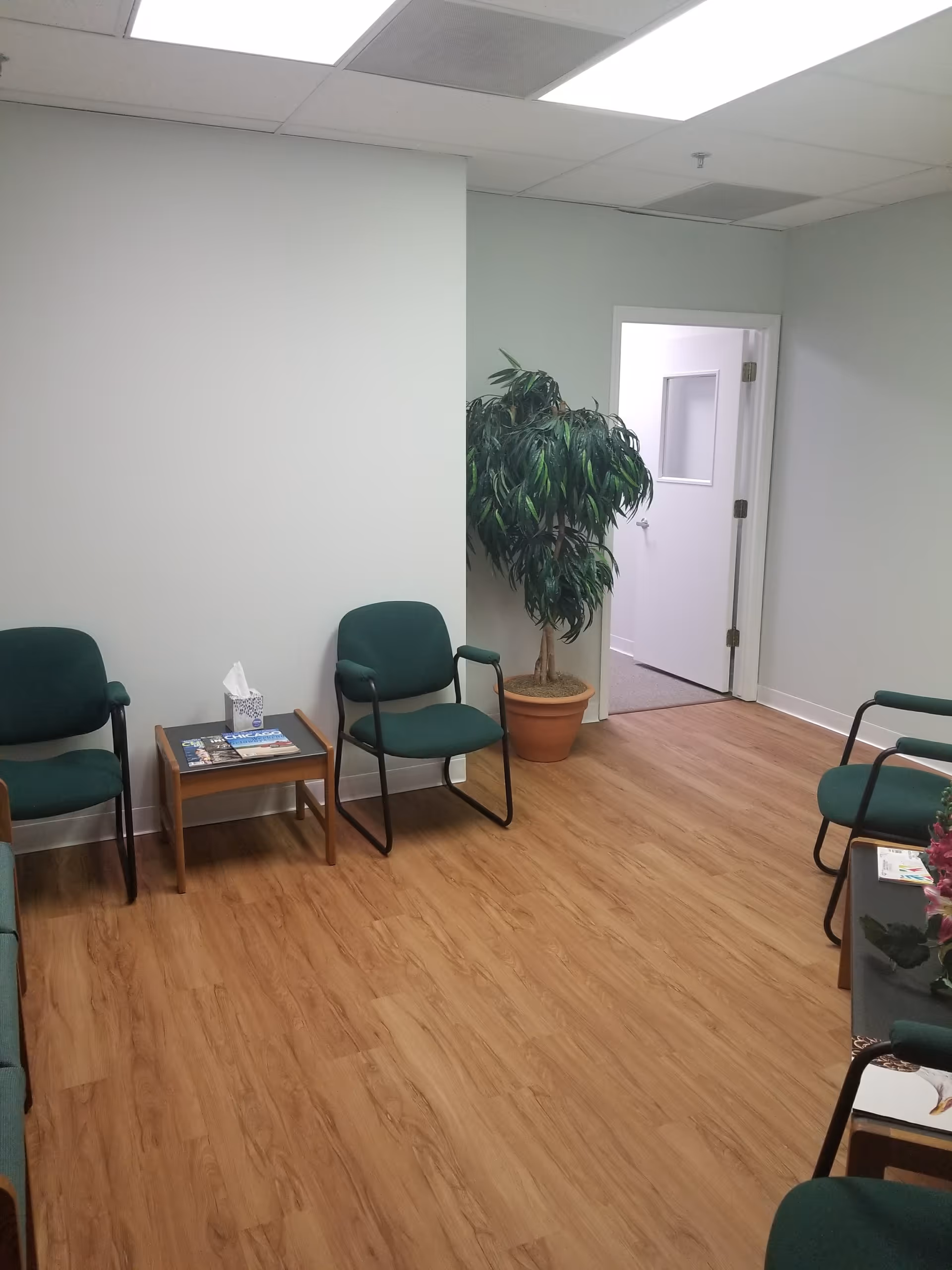 Interior waiting area with green upholstered chairs, small tables, and a potted plant.