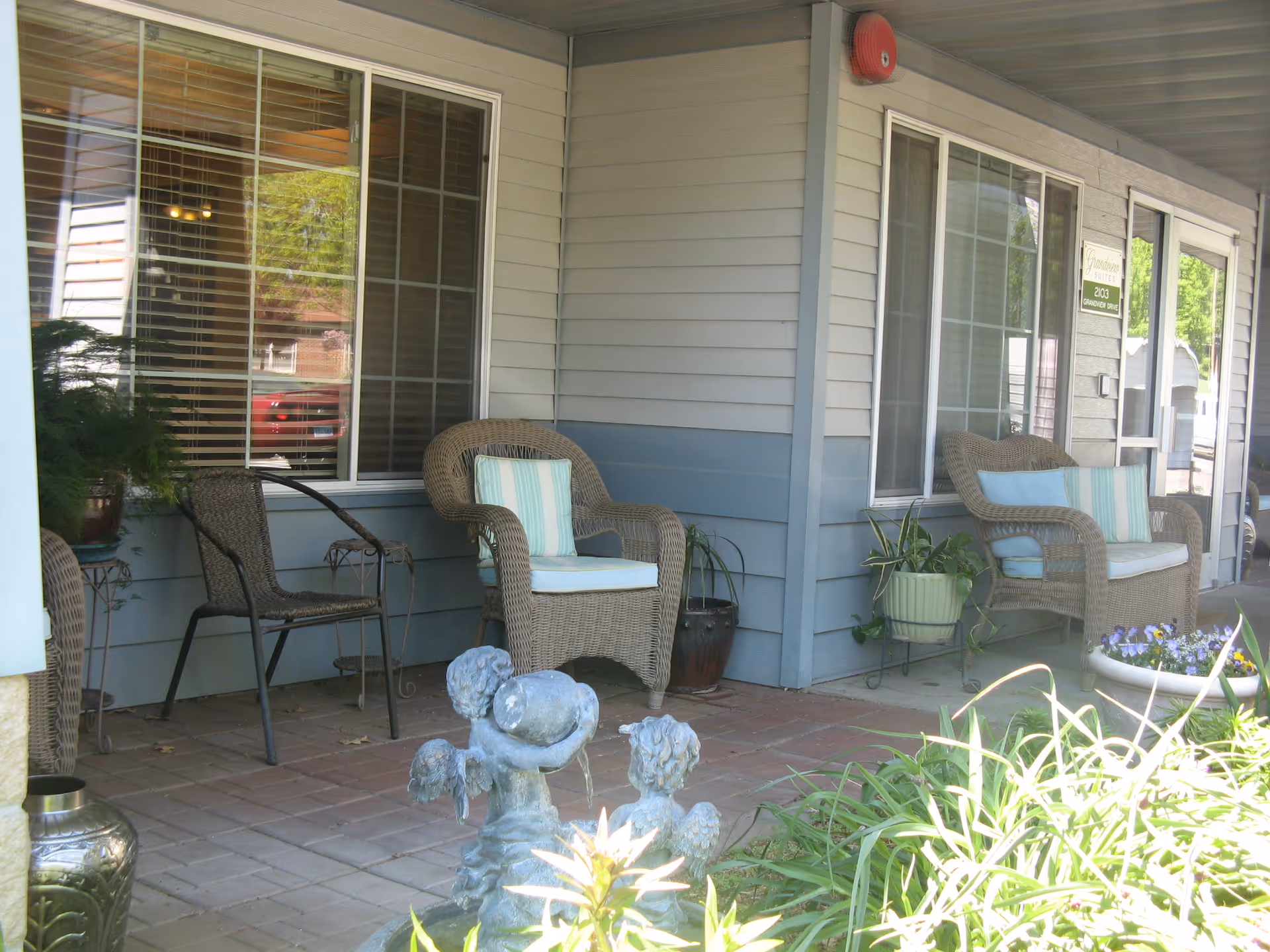 Covered front porch of a senior living facility with wicker chairs, potted plants, and a small fountain.