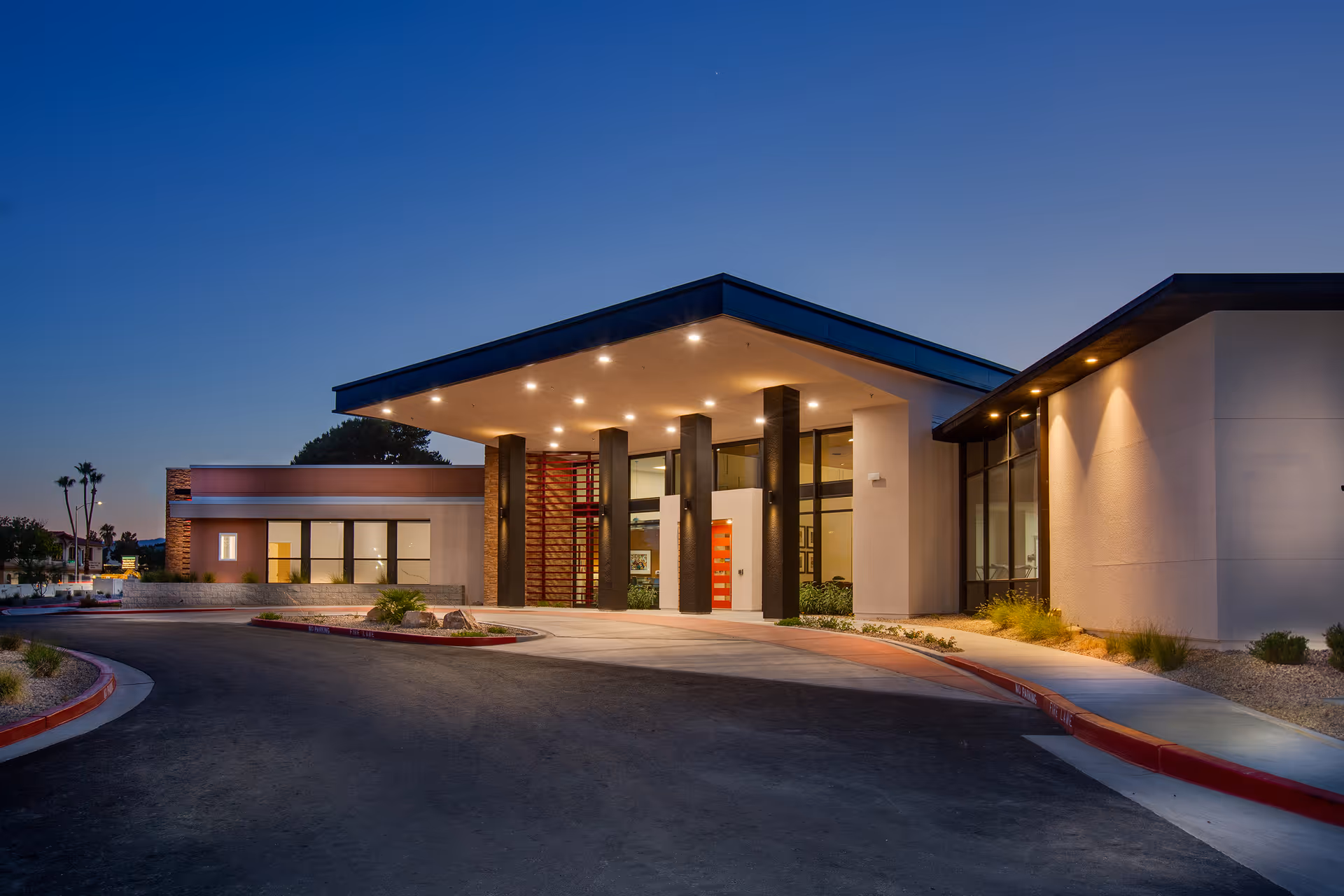 Modern senior living facility with illuminated entrance canopy and curved driveway at dusk.