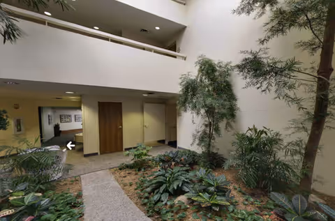 Indoor courtyard area with a variety of green plants and small trees. The space is surrounded by beige walls with a second-floor balcony visible above. There are wooden doors and a hallway leading to other parts of the building.