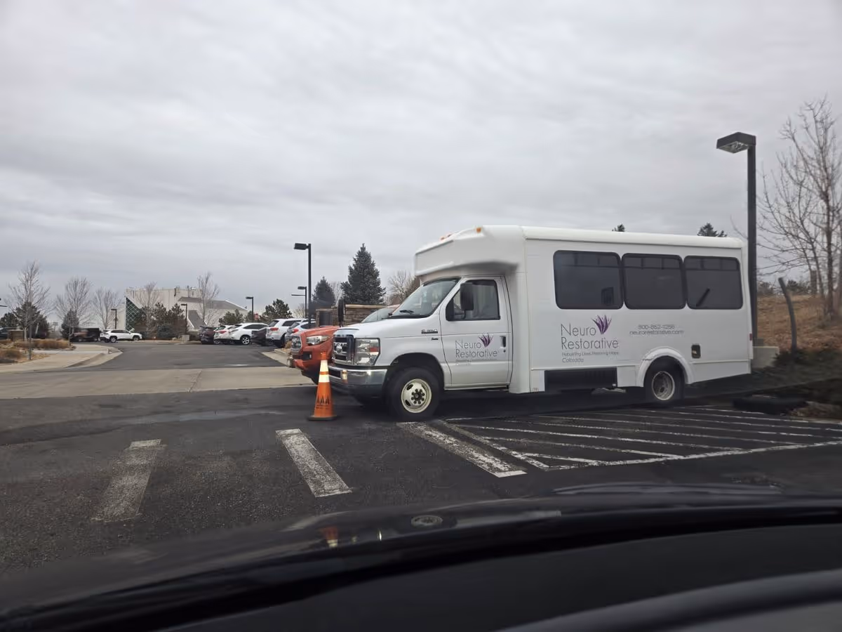 White Neuro Restorative shuttle van parked in an outdoor parking lot in front of a senior living campus under a cloudy sky.