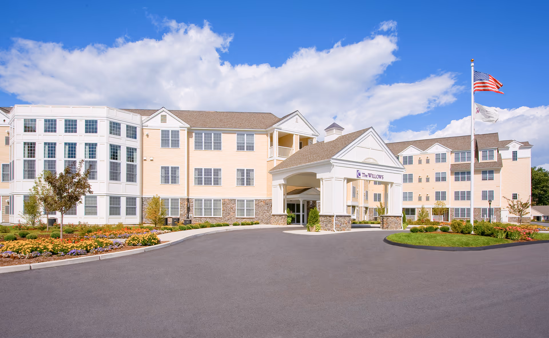 Front exterior view of The Willows Retirement Community building with a covered entrance, landscaped flower beds, and an American flag on a flagpole under a partly cloudy blue sky.
