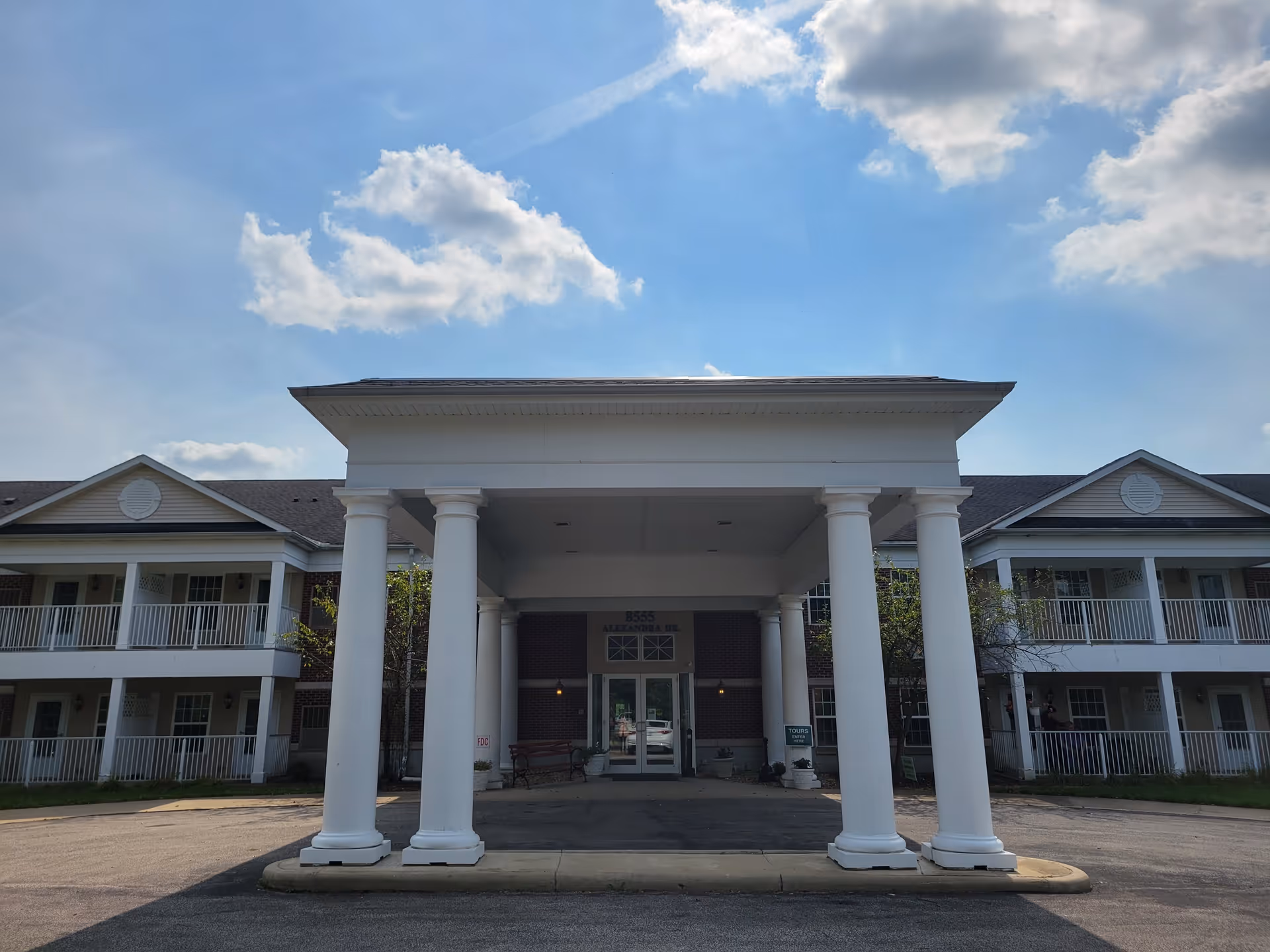 Front exterior view of a two-story building with white columns supporting a covered entrance. The building has balconies with white railings on both sides and a clear blue sky with some clouds above.