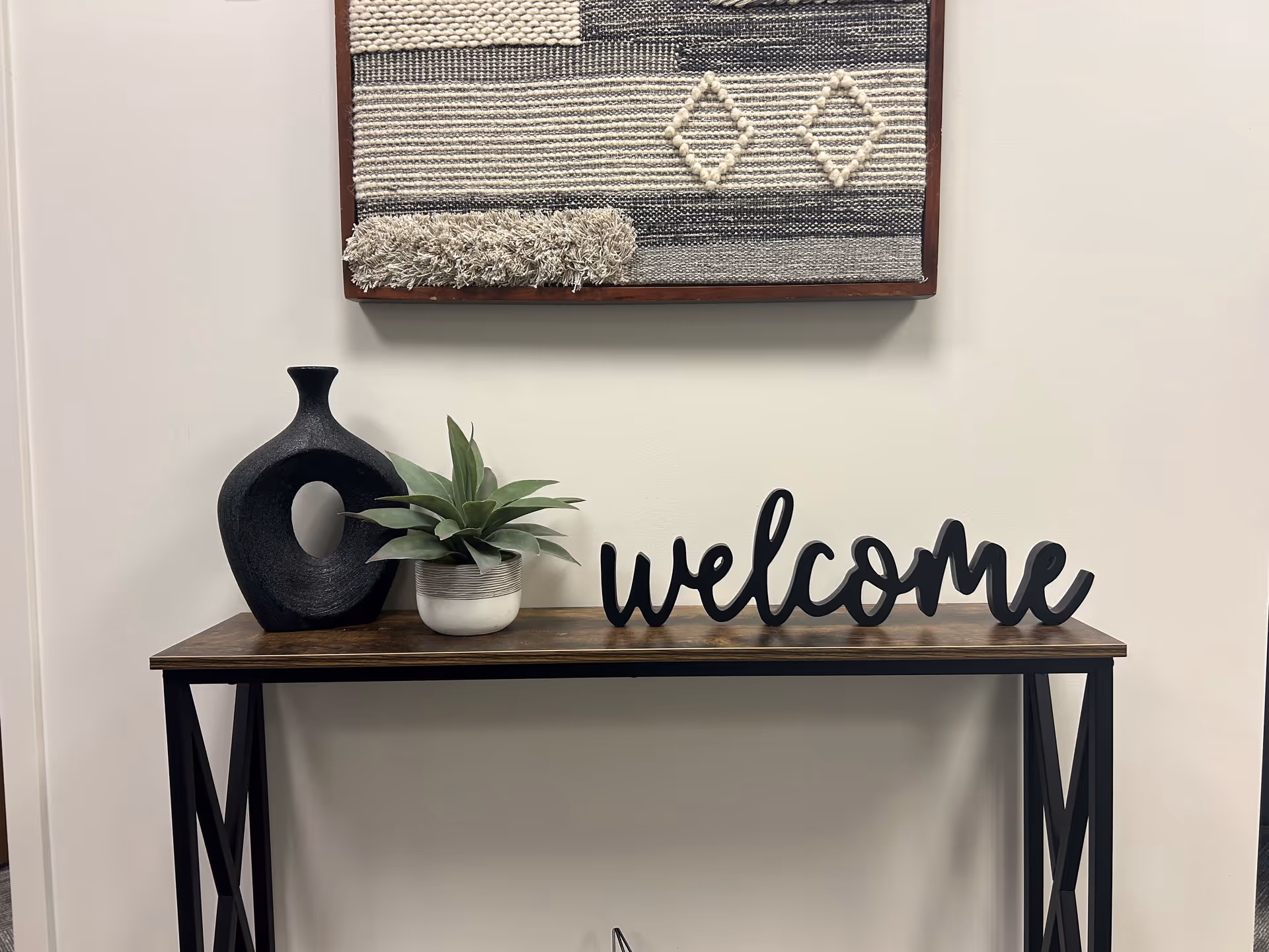 Entryway table with a decorative welcome sign, plant, and ceramic vase against a wall.