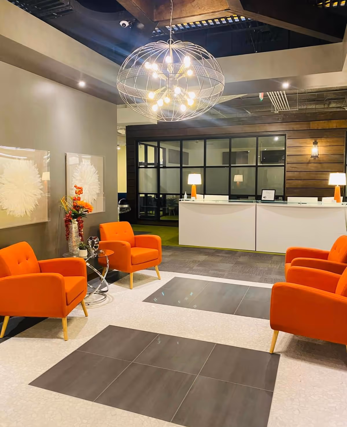 Modern interior lobby with orange armchairs, a circular chandelier, and a glass-partition reception area.