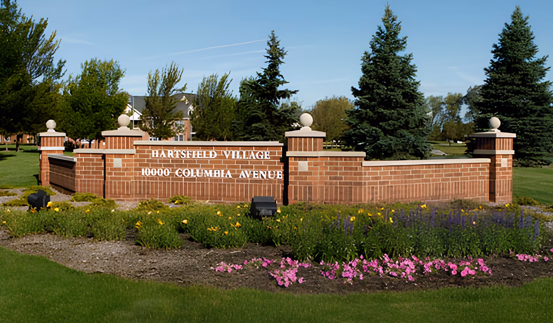 A brick entrance sign for Hartsfield Village with the address 10000 Columbia Avenue, surrounded by green grass, colorful flower beds, and tall trees under a clear blue sky.
