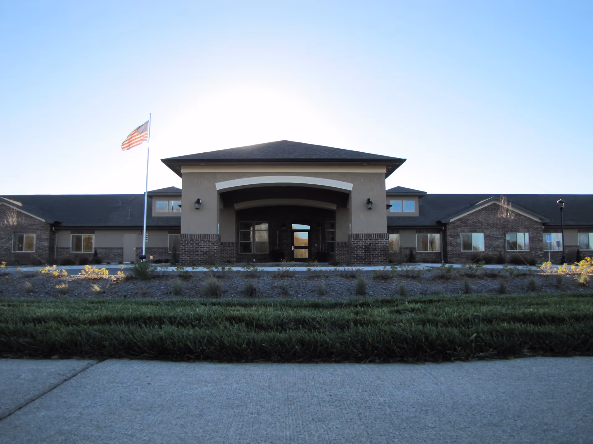 Front exterior of a single-story senior living facility with a covered entrance, flagpole, and landscaped lawn.