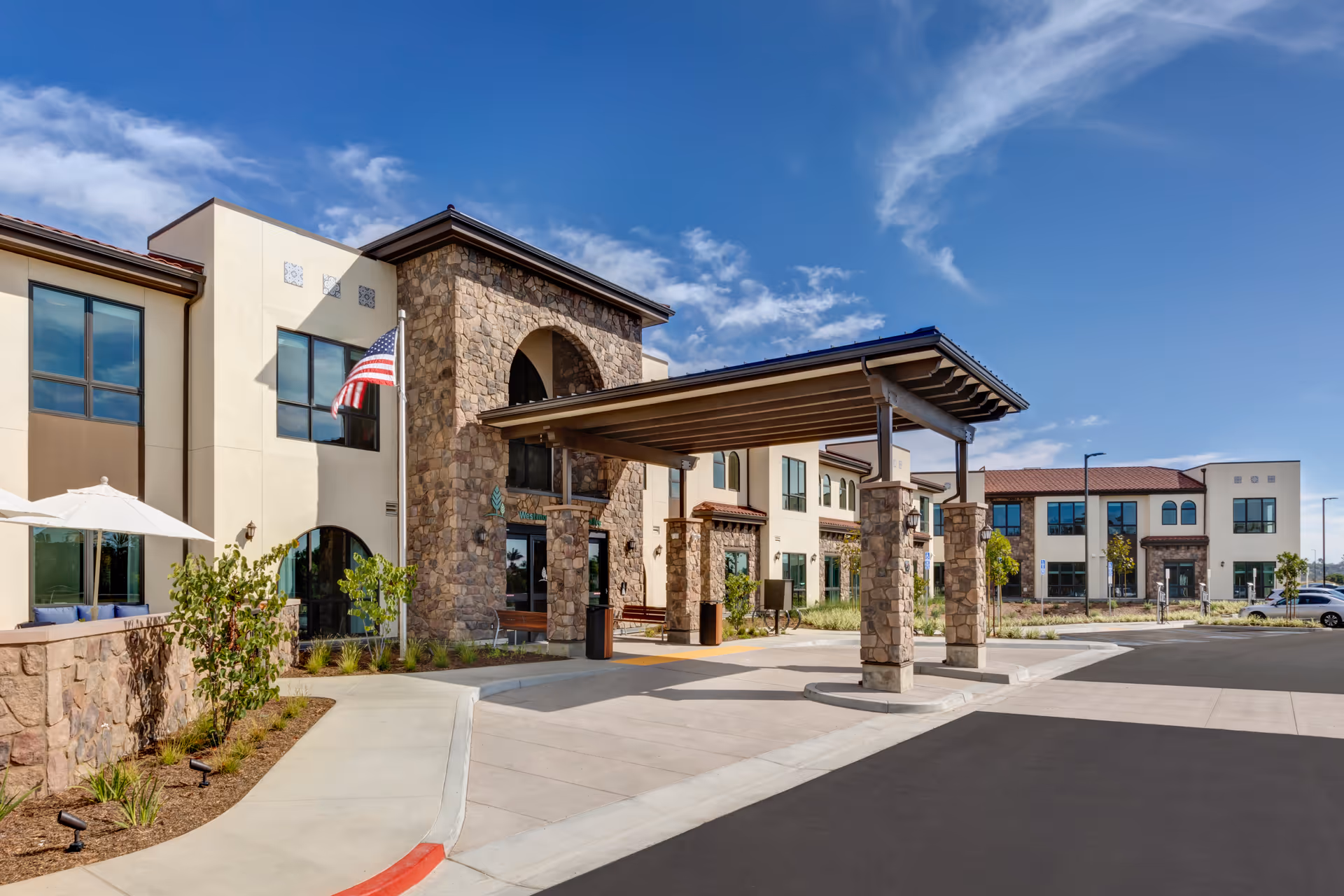 Exterior view of Westmont of Carmel Valley senior living facility showing a two-story building with stone and stucco facade, a covered entrance with stone pillars, an American flag, and a clear blue sky.