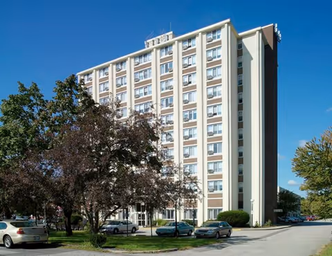Multi-story beige apartment building with parked cars and trees in front under a clear blue sky.