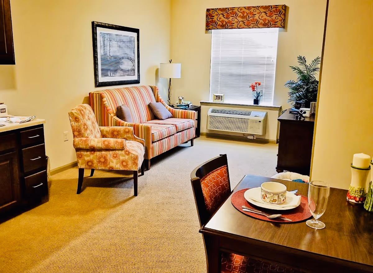 A cozy living area with a striped loveseat and a floral armchair, a window with blinds and a floral valance, a side table with a lamp and decorative items, a wooden dining table set with a bowl, plate, napkin, and glass, and a cabinet with a plant and other decor.