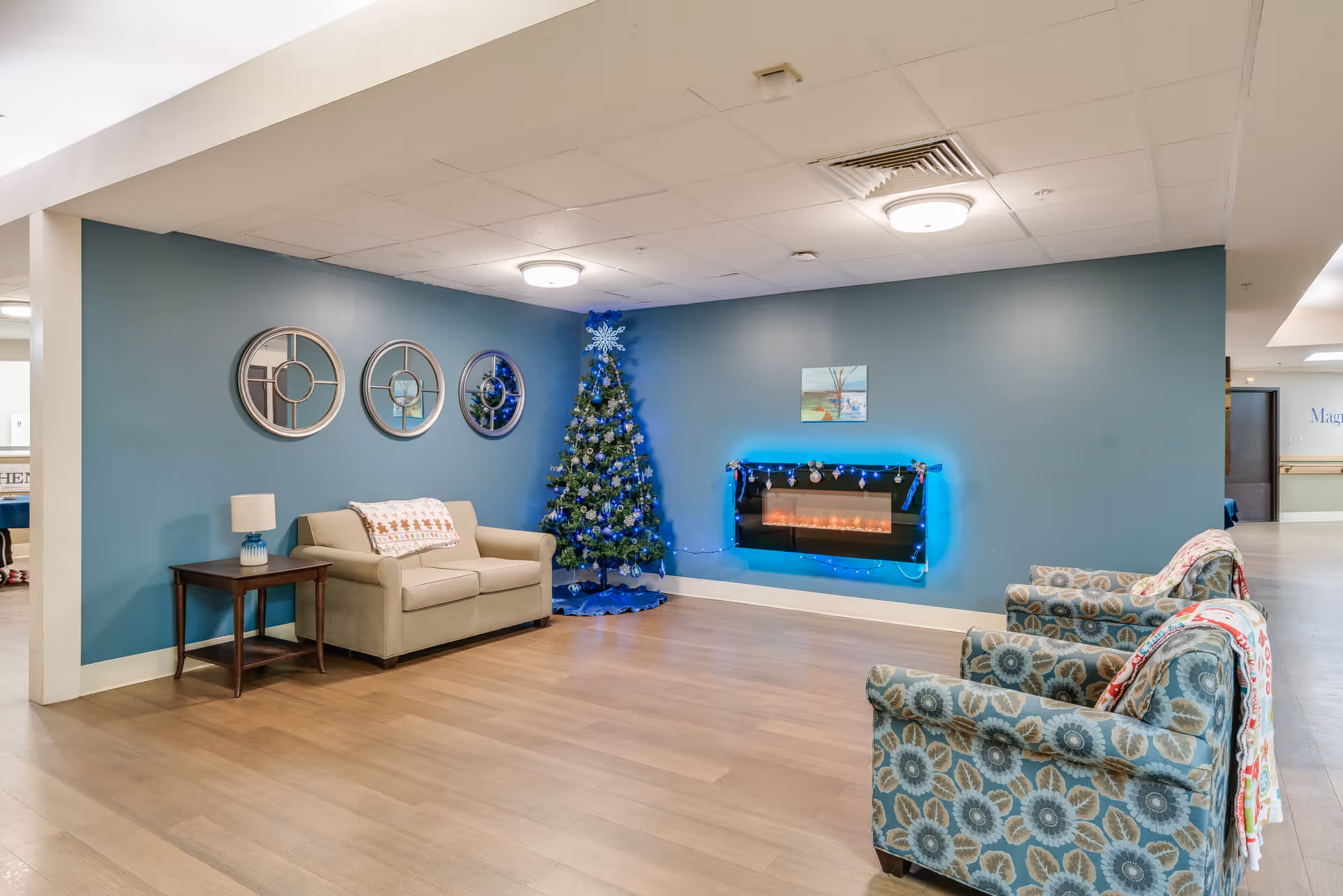 A cozy senior living common area with a beige loveseat next to a wooden side table with a lamp, two patterned armchairs with blankets draped over them, a decorated Christmas tree, and a modern electric fireplace with blue accent lighting mounted on a blue wall. Three round mirrors hang on the wall above the loveseat.