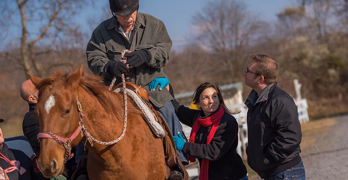Outdoor horse ride with staff guiding a rider and onlookers.
