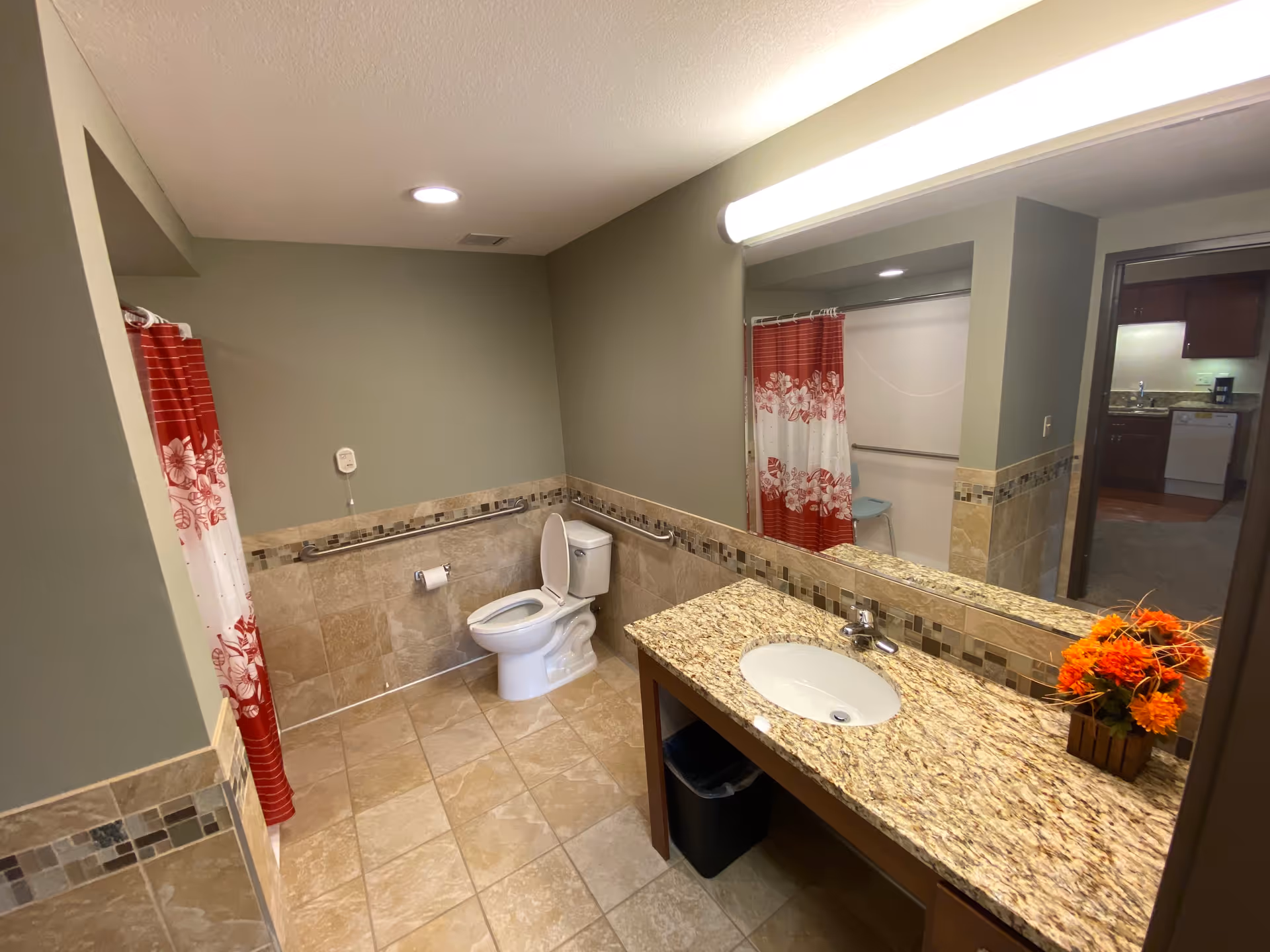 A spacious bathroom with beige tiled floor and walls partially tiled with decorative mosaic trim. There is a white toilet with a grab bar beside it, a shower area with a red and white floral shower curtain, and a granite countertop with an oval sink. A large mirror spans the wall above the sink, reflecting the shower and part of an adjacent room. A small vase with orange flowers is placed on the countertop.