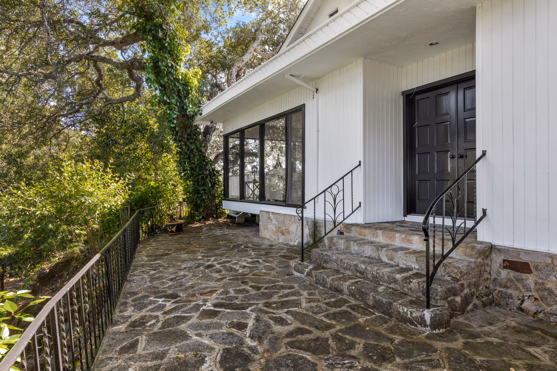 Stone patio area outside a white building with black double doors and black metal railings on the steps. The patio is surrounded by trees and greenery, with a black metal fence along the edge.