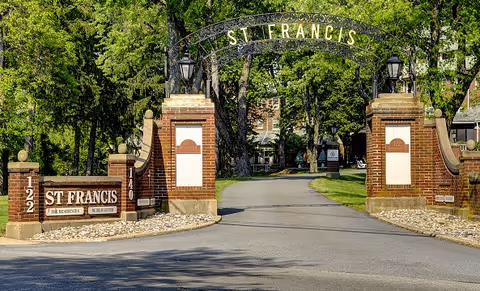 Stone gate arch reads Saint Francis Residential Community, brick pillars frame a tree-lined driveway.