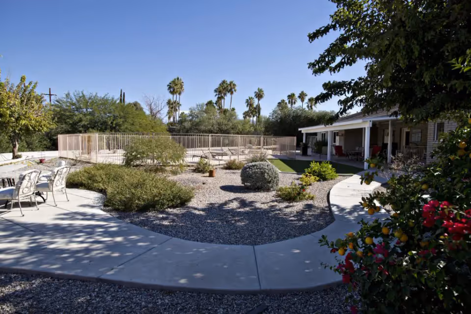 Sunny courtyard with gravel landscaping, paved walkways, patio seating, a fenced pool, and a single-story building with a covered porch and palm trees in the background.
