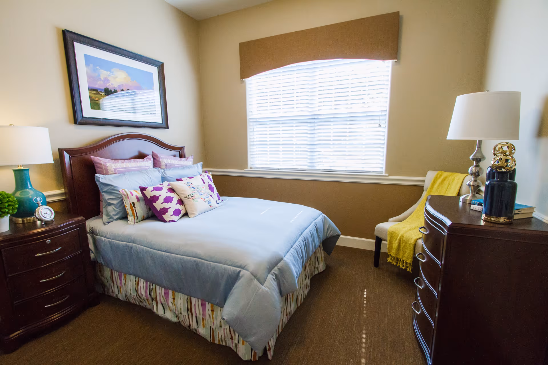 A cozy bedroom with a neatly made bed featuring light blue bedding and multiple decorative pillows. There is a wooden nightstand with a blue lamp and a small plant on the left side of the bed. A framed landscape painting hangs on the wall above the headboard. A window with white blinds and a brown valance is centered on the back wall. On the right side, there is a wooden dresser with a white lamp, decorative items, and a chair draped with a yellow blanket.