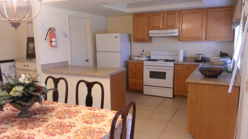 Kitchen area with wooden cabinets, stove, fridge, and a floral-draped dining table in foreground.