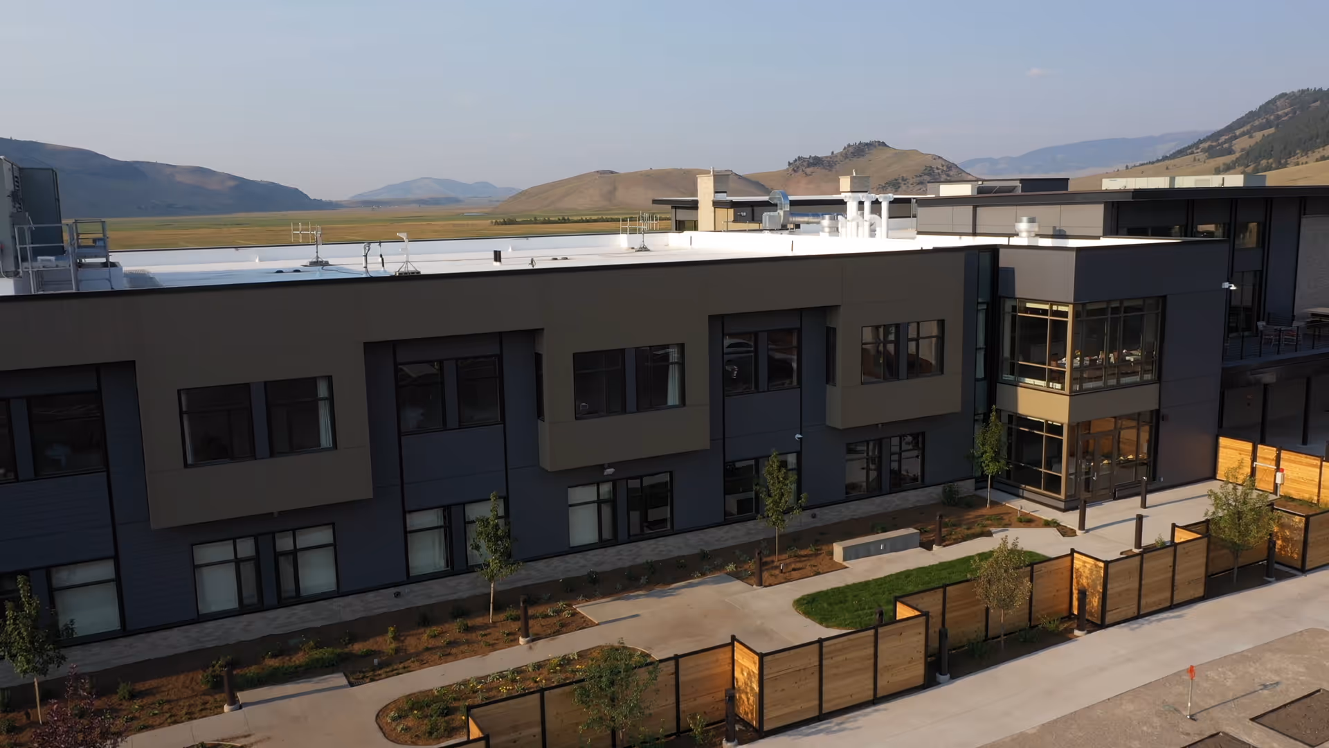 A modern two-story senior living center exterior with balconies, walkways, and surrounding hills.