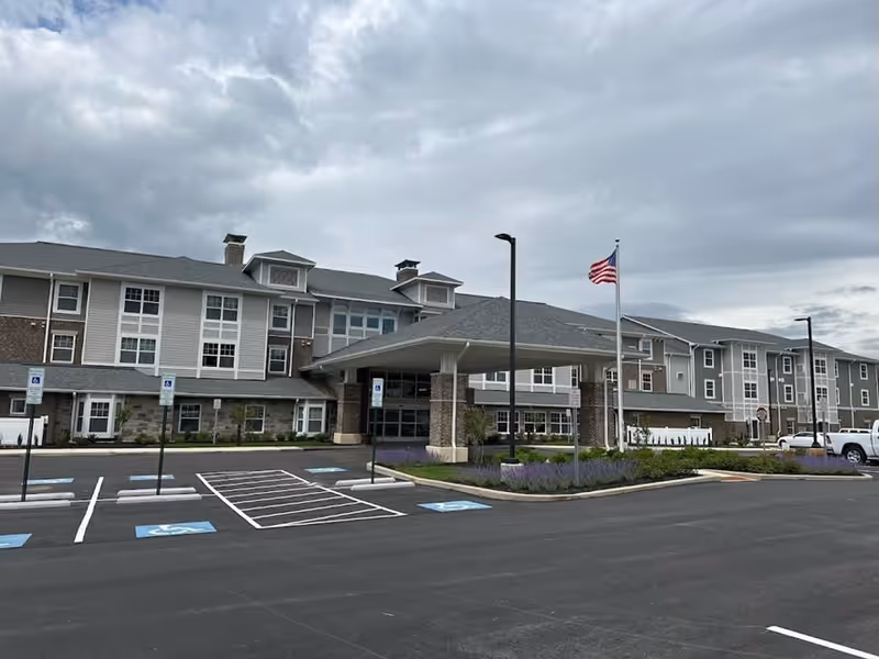 Exterior view of a large, multi-story retirement community building with a covered entrance, several windows, an American flag on a flagpole, and a parking lot with handicap parking spaces in front under a cloudy sky.